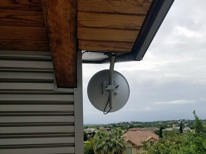 Dish on a bent-pipe mount under a wooden eave, aimed across a Hill Country neighborhood