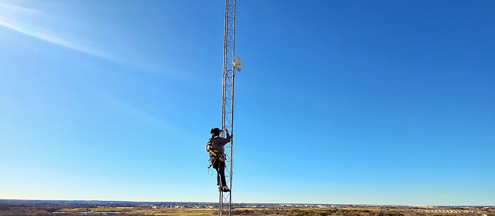 Engineer Austin climber in a fall-protection harness partway up a lattice tower with point-to-point dishes, high above a river valley