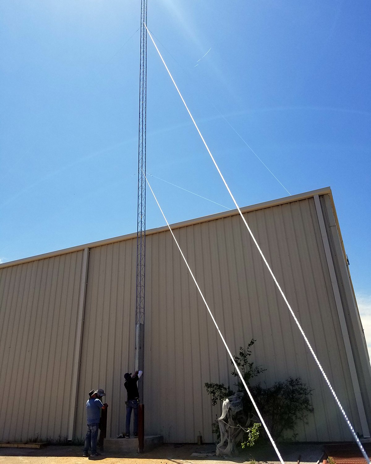 Engineer Austin crew raising a guyed lattice tower beside a metal building, guy wires running to the ground