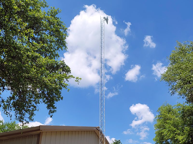 Lattice tower bracketed to the eave of a metal shop building, with a dish at the top and live oaks around it